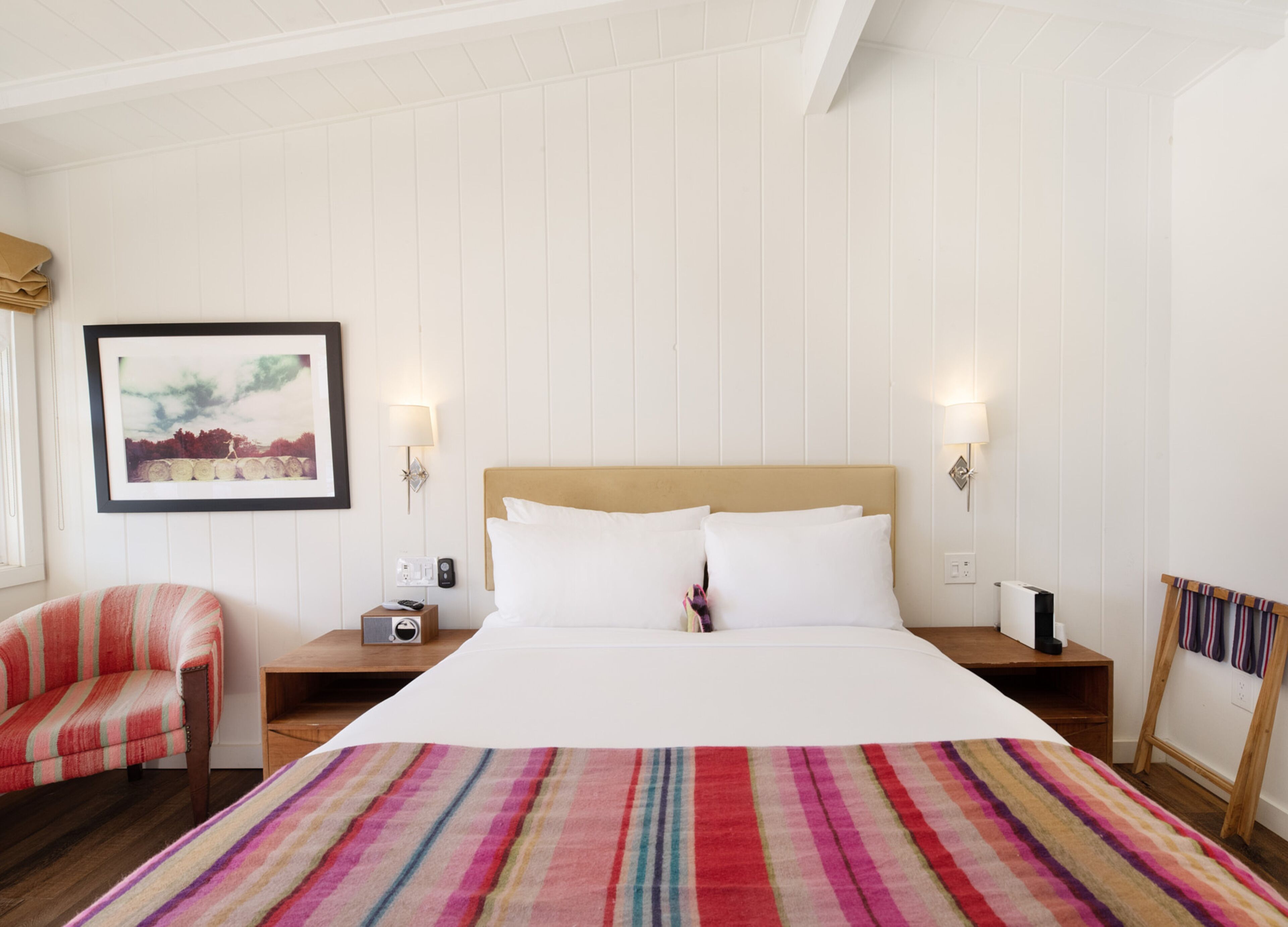 Ein Hotelzimmer mit einem großen Bett, zwei Nachttischen, einem Sessel, einem gerahmten Bild an der Wand und einer gestreiften Decke.
