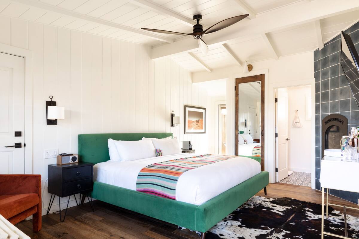 Quarto com cama estofada verde, roupa de cama branca, mesa de cabeceira de madeira, rádio, abajur preto, tapete de couro de vaca, banco de madeira, ventilador de teto e banheiro com espelho e artigos de higiene pessoal.