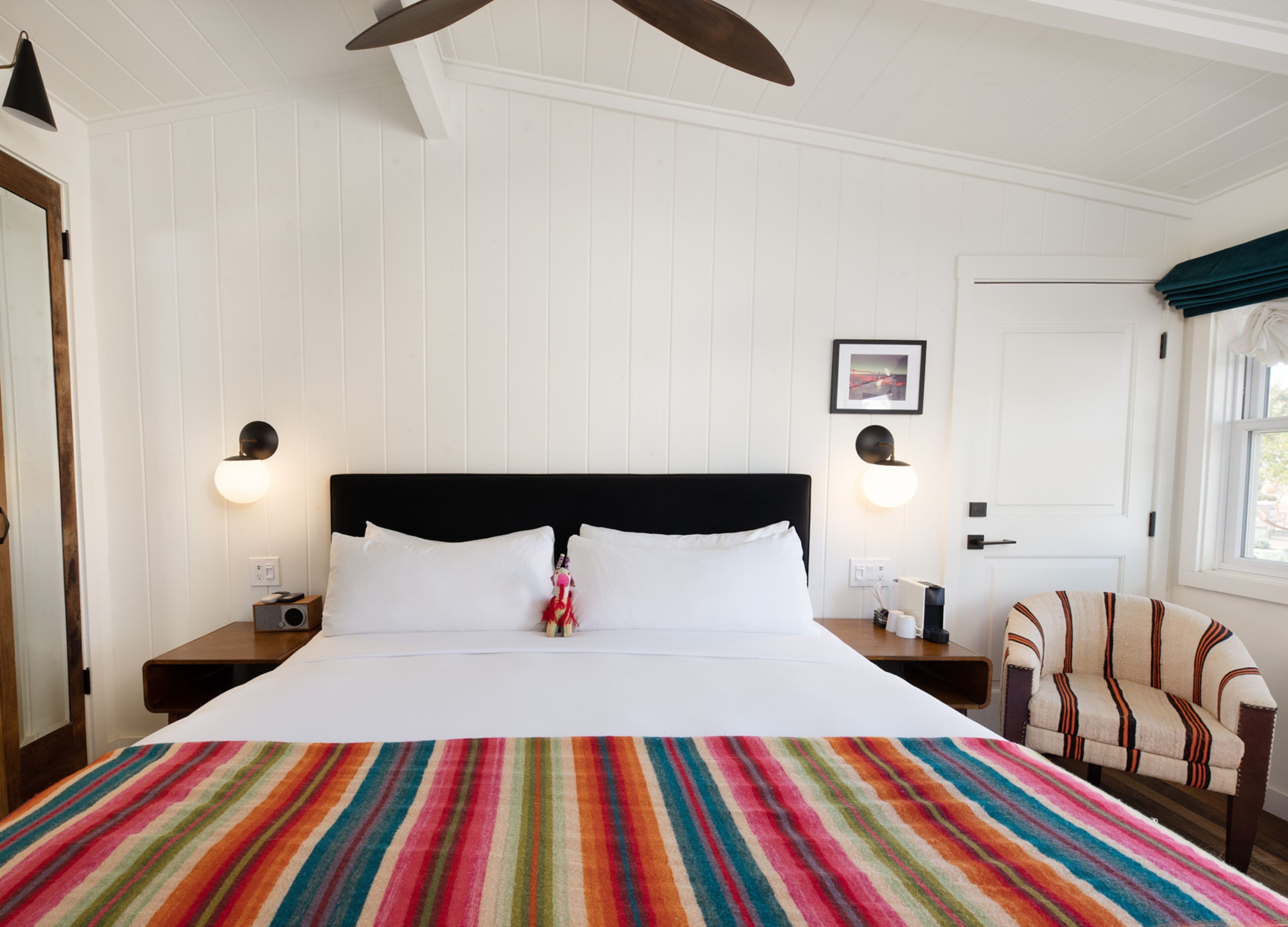 Habitación de hotel con una cama grande, dos mesitas de noche, una silla, un ventilador de techo y una puerta con ventana.