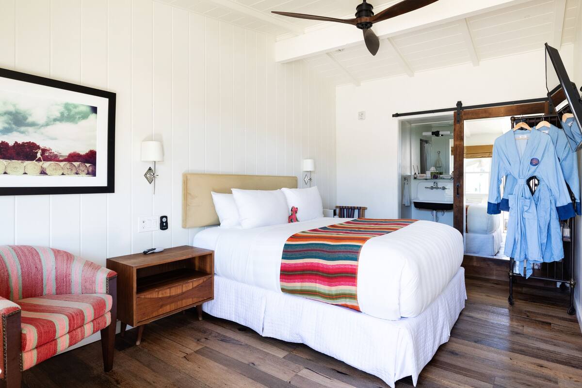 Quarto de hotel com uma cama grande, uma mesa de cabeceira de madeira, uma cadeira, um ventilador de teto e um banheiro visível através de uma porta aberta.