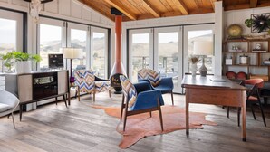 Amplia sala de estar con techo de madera alto, ventanas grandes con vistas a las colinas, chimenea y una mezcla de muebles modernos y rústicos.