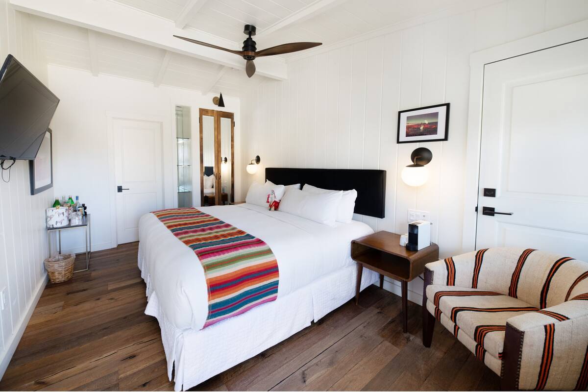 Quarto de hotel com uma cama grande, um ventilador de teto, uma mesa de cabeceira com abajur, uma cadeira e uma televisão fixada na parede.