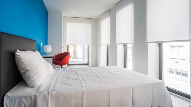 Un dormitorio moderno con una cama grande, una silla roja y vistas de la ciudad a través de amplias ventanas.