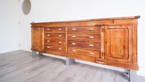 Un aparador de madera con varios cajones y espacios de almacenamiento en una habitación con suelo de madera clara y paredes blancas.