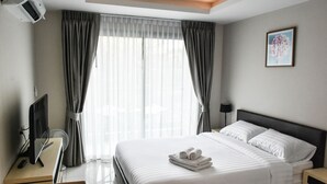 A hotel room with a bed, a desk with a computer, a chair, a desk lamp, and a window with curtains. - Water Park Condominium by Able Estate (Pattaya)
