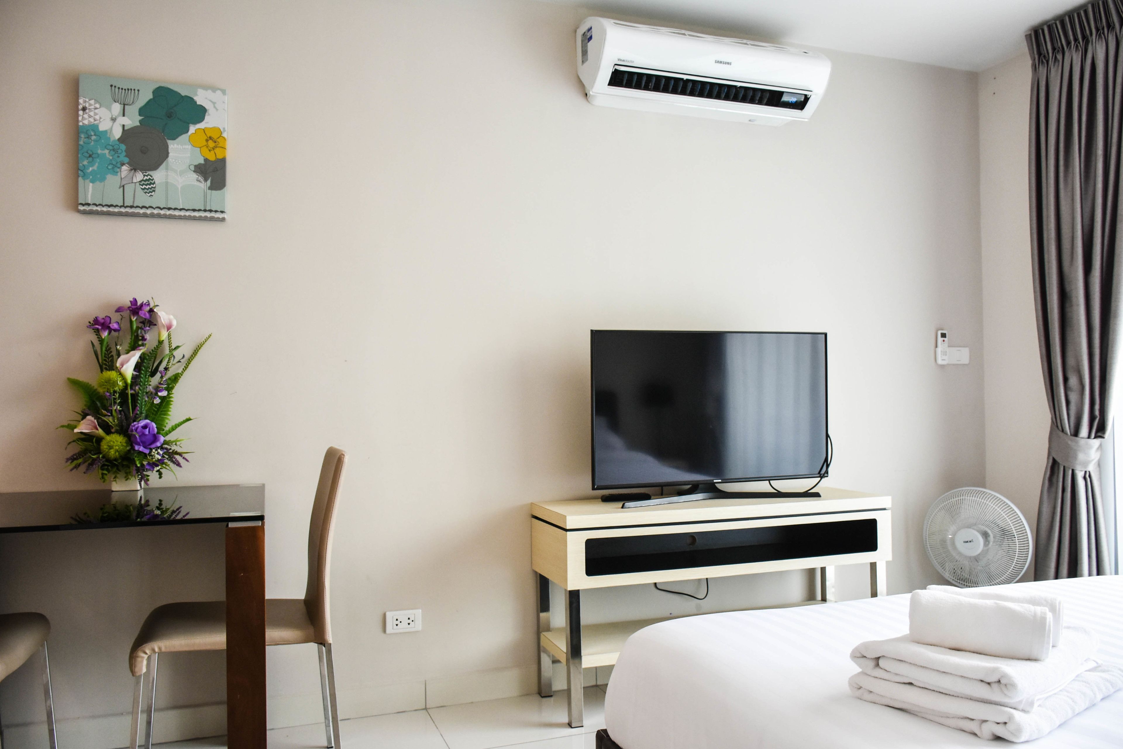 Een hotelkamer met een bed, een televisie op een standaard, een airconditioner, een bureau met een stoel, en een ventilator aan de muur.