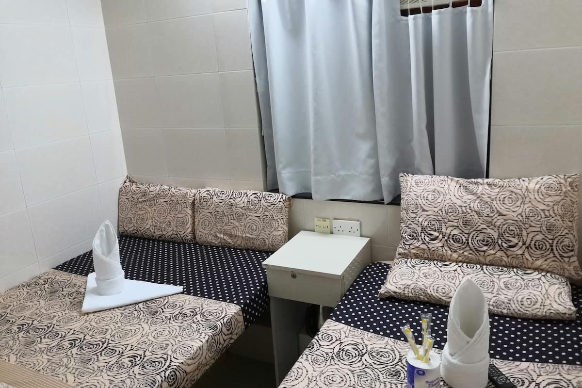 A small, neatly arranged room with a bed, a bedside table, and a chair. There is a window with curtains, a wall-mounted air conditioning unit, and a power outlet.
