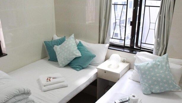 A hotel room with two beds, each with white linens and teal pillows. There is a small bedside table with a remote control, a tissue box, and a cup. The room has a window with curtains, and the walls are tiled. - New Yan Yan Guest House (Kowloon)