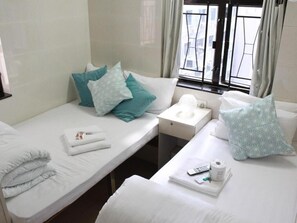 A hotel room with two beds, each with white linens and teal pillows. There is a small bedside table with a remote control, a tissue box, and a cup. The room has a window with curtains, and the walls are tiled. - New Yan Yan Guest House (Kowloon)