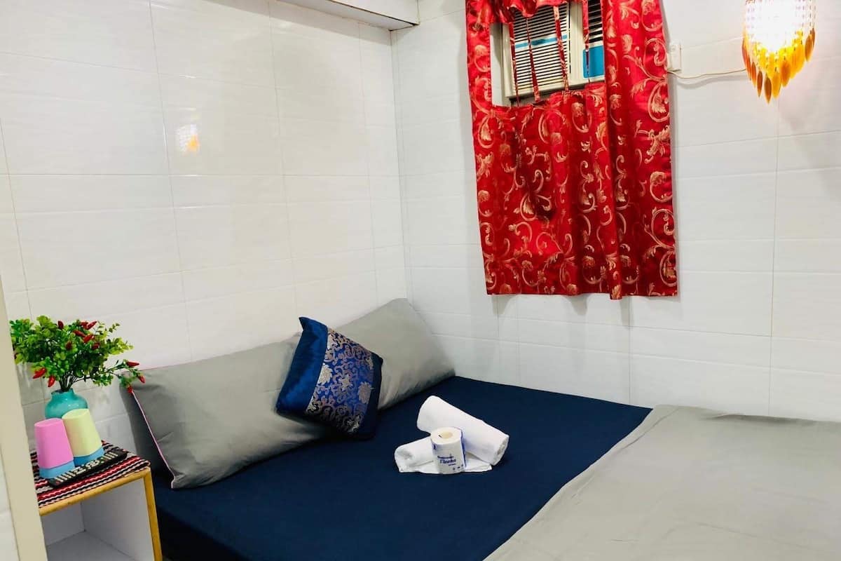A small, neatly arranged room with a bed, a shelf with toiletries, a window with red curtains, and a wall-mounted light fixture.