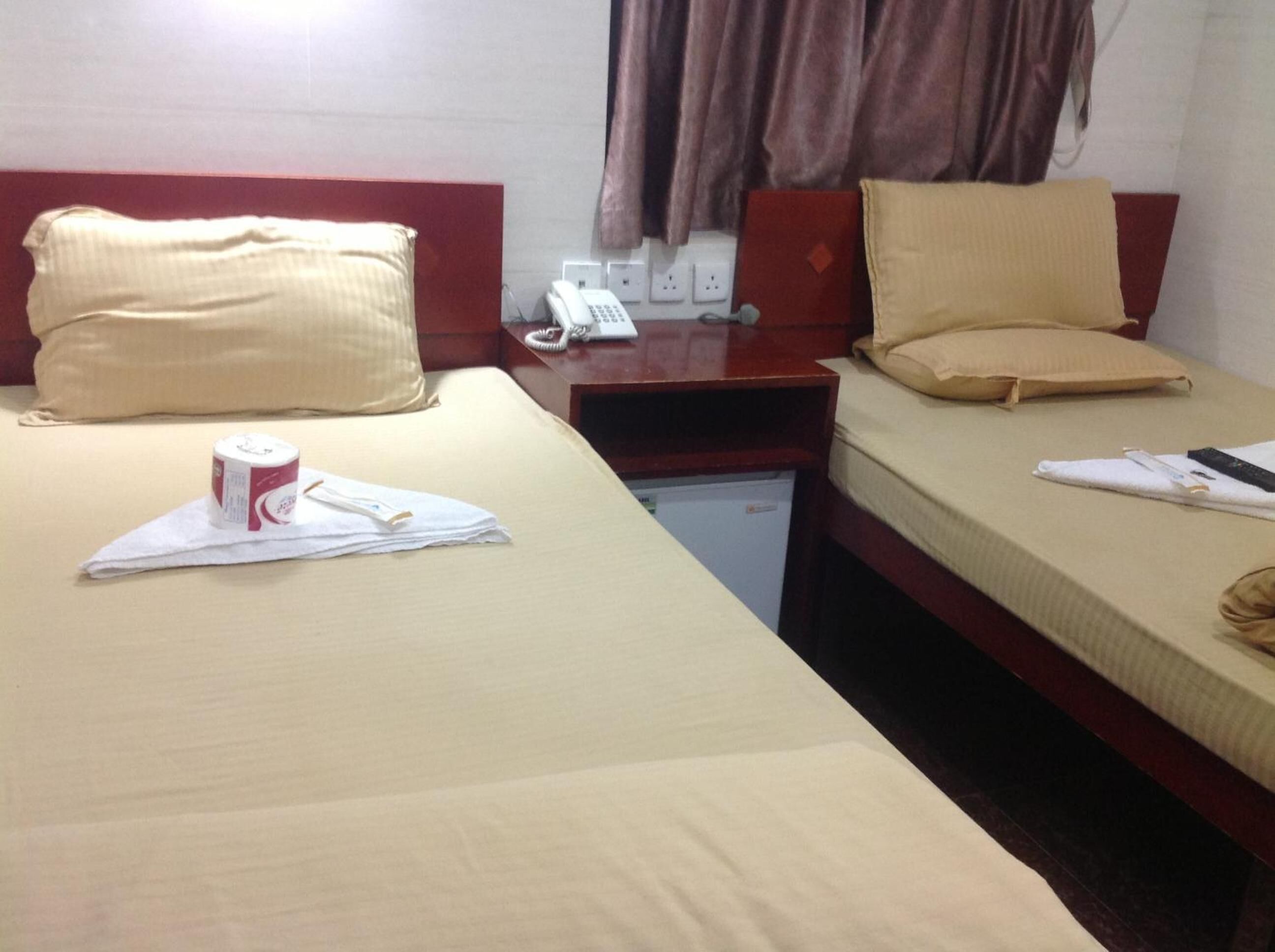 A hotel room with two beds, each with beige bedding and pillows. There is a small desk with a telephone and a red folder. A white towel and a pair of glasses are on the left bed.
