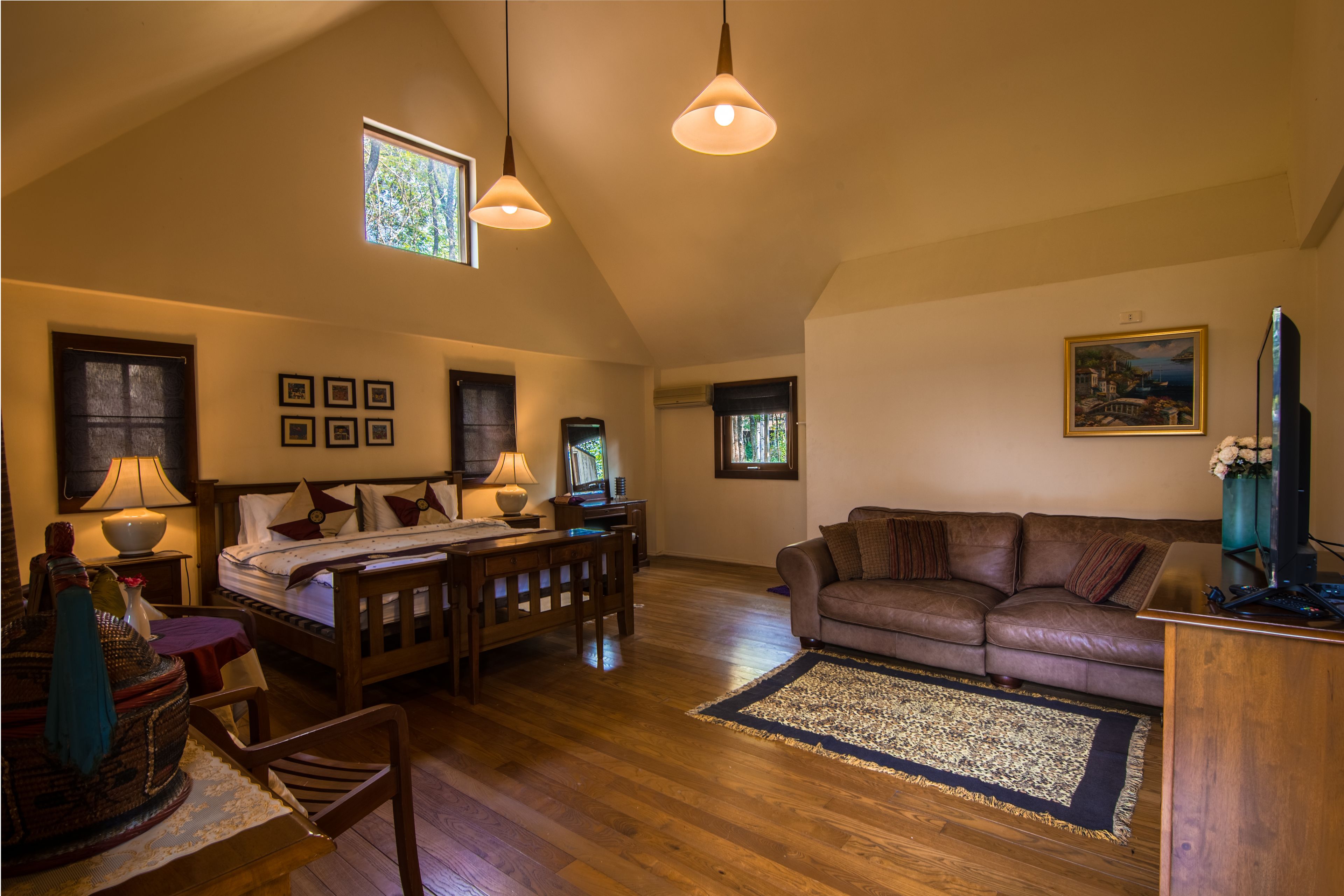 A spacious living room with a wooden floor, a brown leather sofa, a dining table with chairs, a bed with a wooden headboard, and a large window providing natural light.