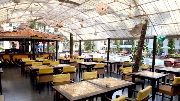 A covered dining area with tables and chairs, a bar, and a pool in the background. - Alor Grande Holiday Resort (Candolim)