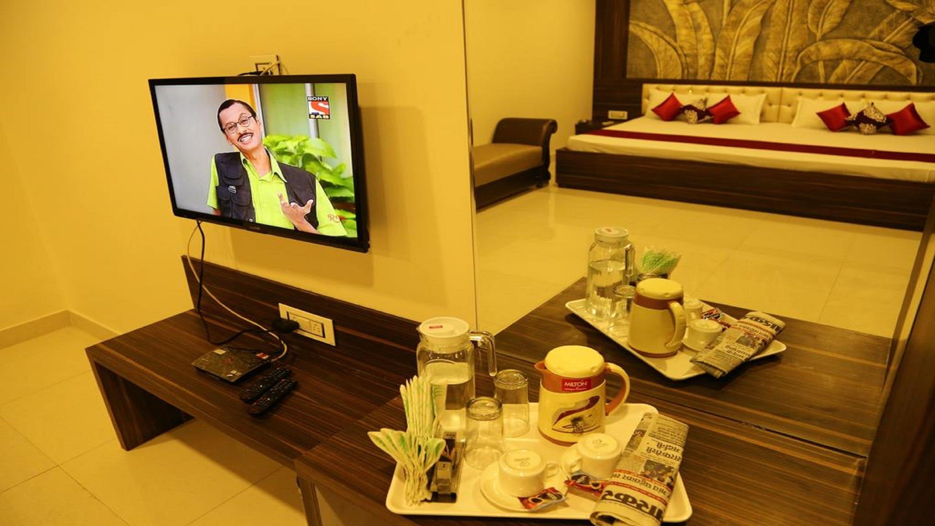 A hotel room with a television displaying a news program, a bedside table with a tray of breakfast items, and a bed with red pillows.