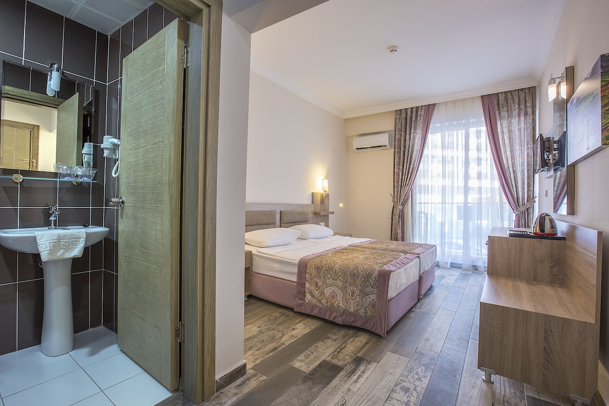 Une chambre d’hôtel avec un lit, une table de chevet, un miroir, un lavabo et une fenêtre avec des rideaux.