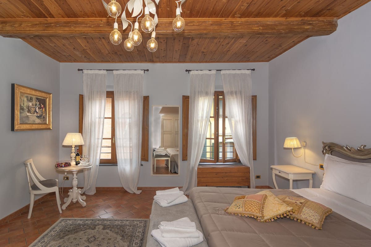 A bedroom with a wooden ceiling, a bed with pillows, a nightstand, a chair, a table with a lamp, and a window with curtains.