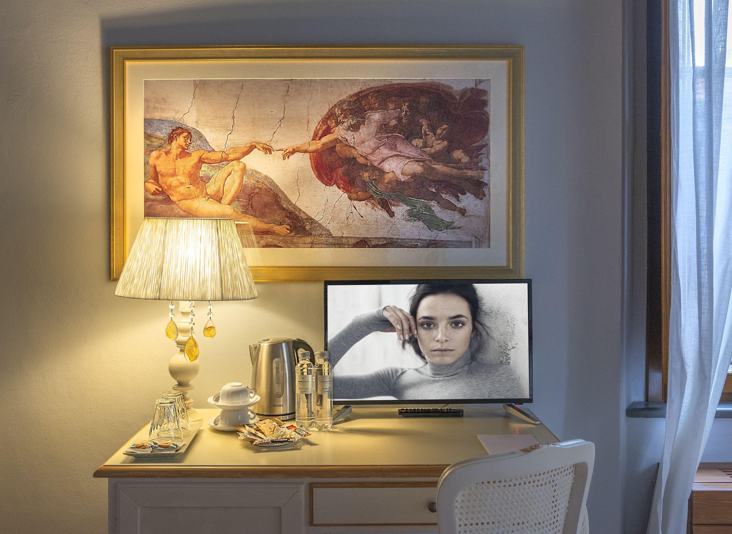A hotel room with a desk, a framed painting, a television, and a lamp.