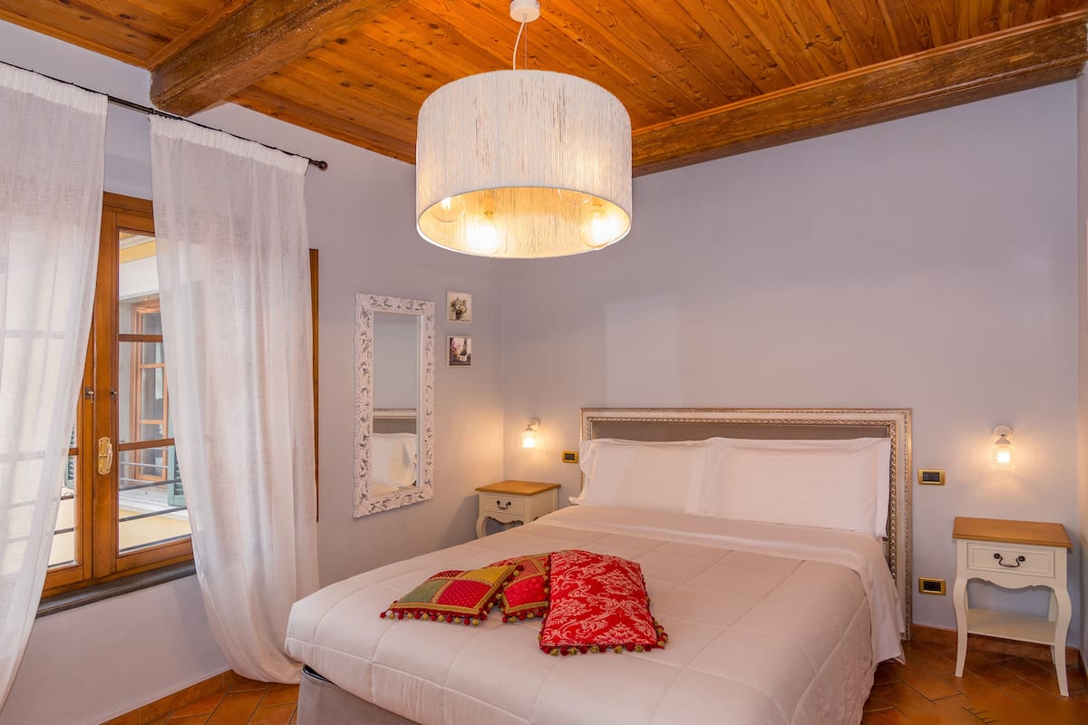 A bedroom with a wooden ceiling, a bed with white bedding and red pillows, a hanging light fixture, and a window with curtains.
