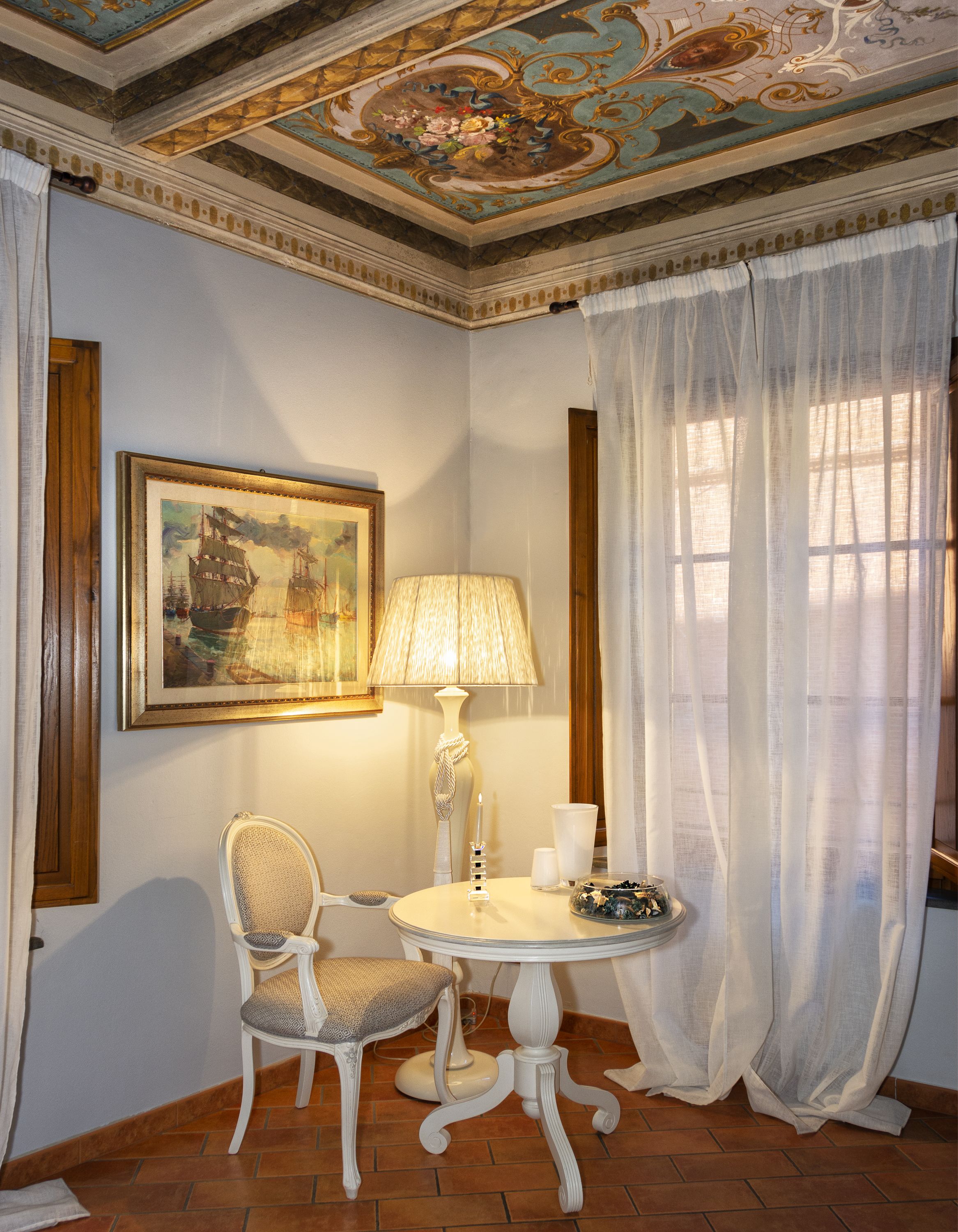 A corner room with a white chair, a table with a lamp, a framed painting, and a window with sheer curtains.