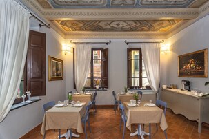 Un comedor con cuatro mesas, cada una con mantel y sillas azules. Hay dos ventanas con cortinas, un cuadro colgado en la pared y un techo con diseños decorativos.