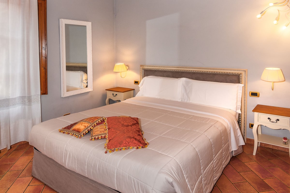 A hotel room with a large bed, two bedside tables with lamps, a mirror, and a decorative ceiling.