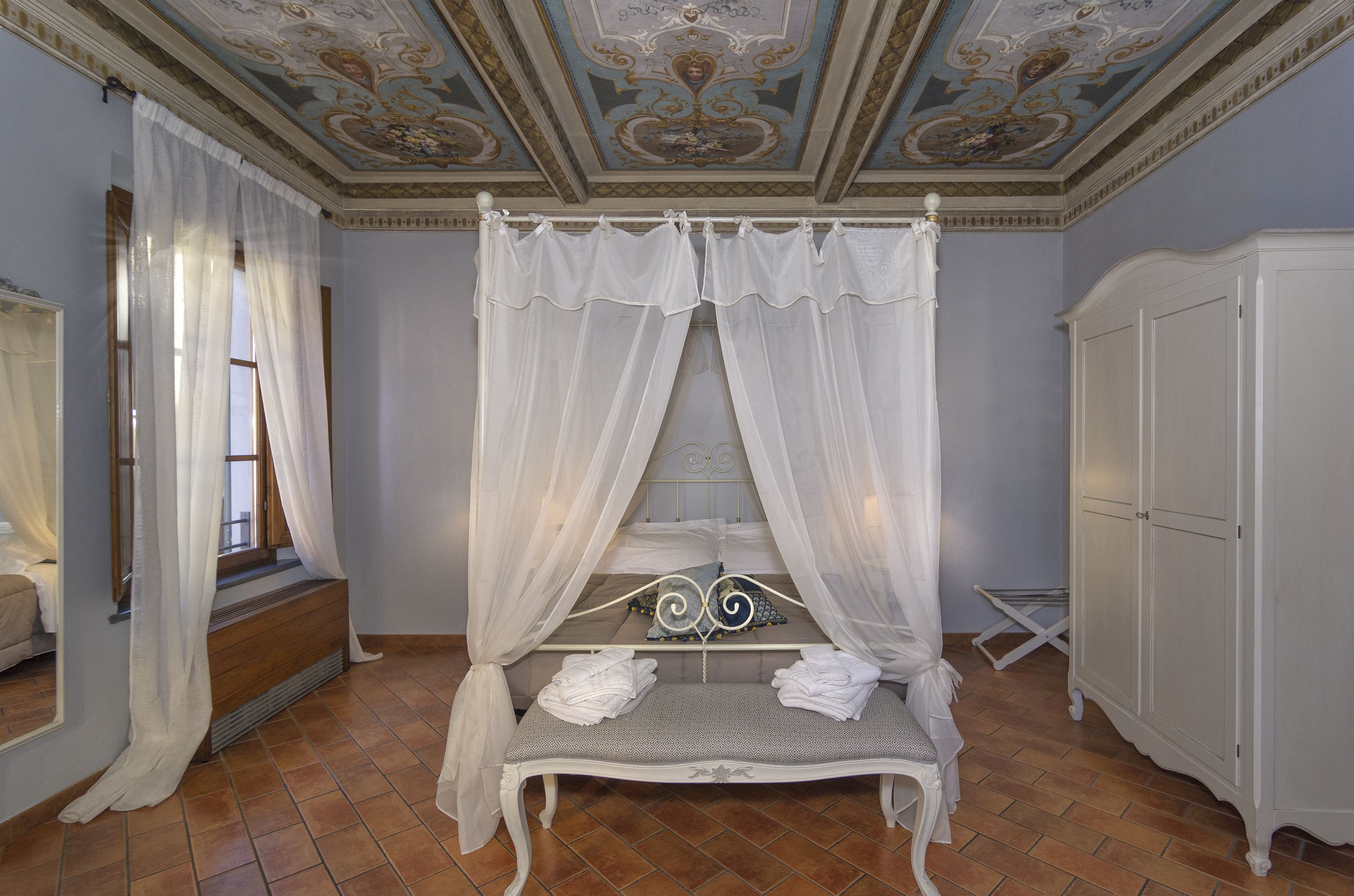 Un dormitorio con cama con dosel, dosel blanco, banco de madera con toallas, armario blanco y techo decorativo.