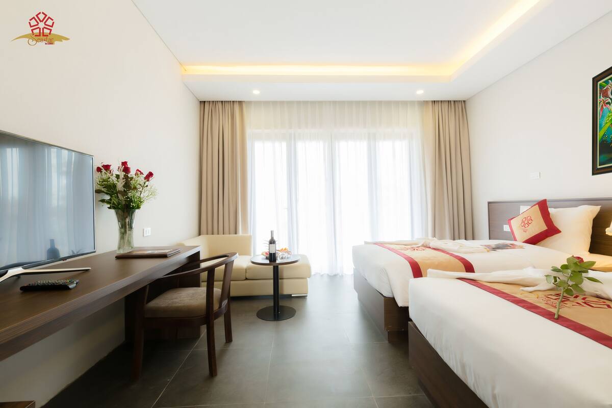 A hotel room with a bed, a desk with a TV, a small table with a vase of flowers, and a sofa.
