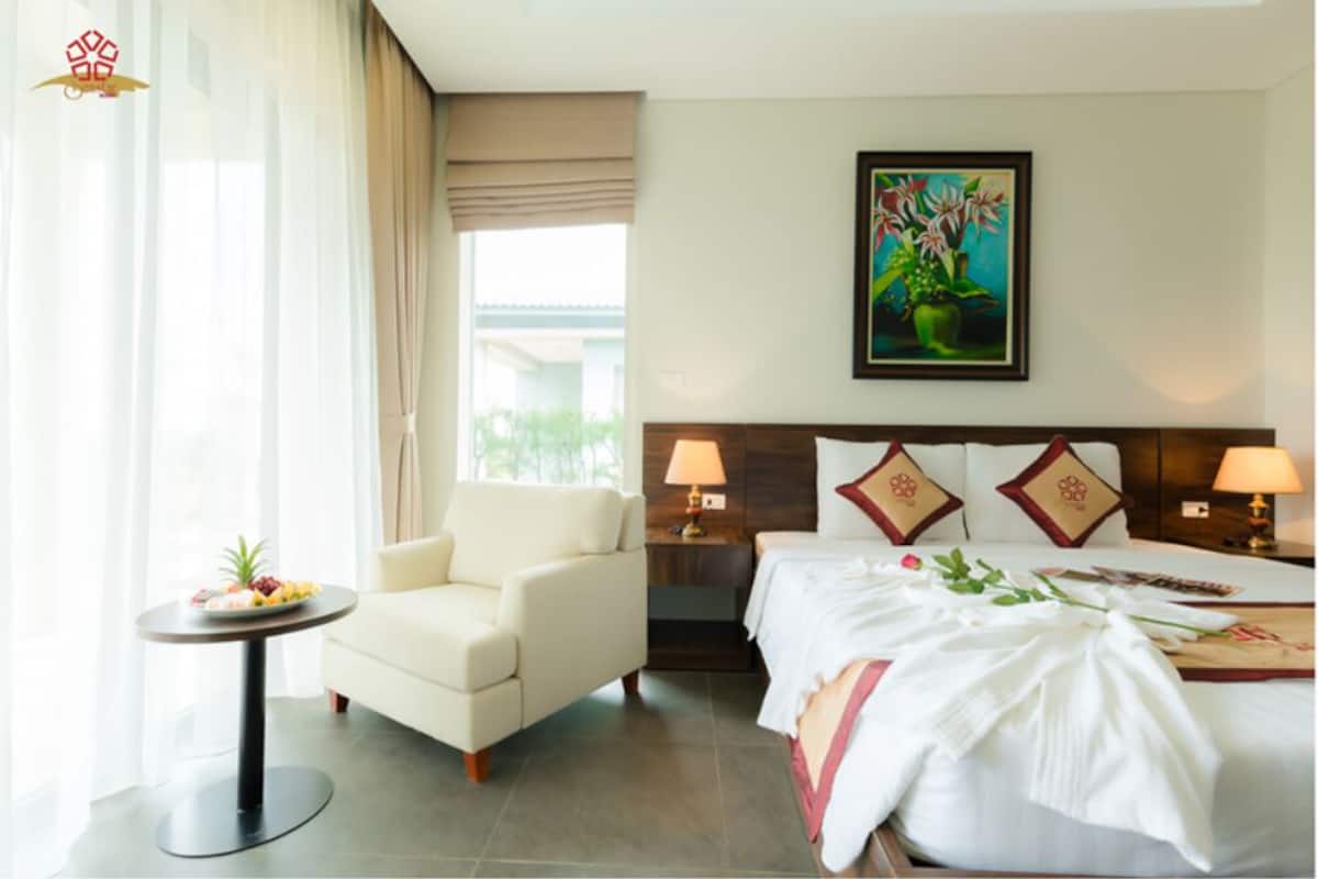 A hotel room with a bed, a chair, a small table with a fruit plate, a painting on the wall, and a window with curtains.