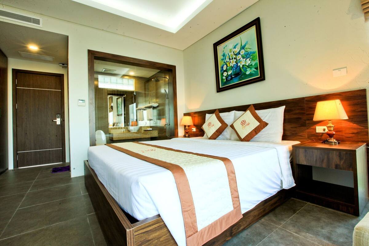 A hotel room with a large bed, a wooden headboard, a nightstand with a lamp, a painting on the wall, and a mirror above the bed.