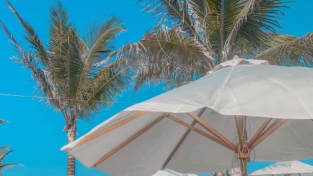 Een vrouw zit op een houten ligstoel onder een grote parasol, met een zwembad en palmbomen op de achtergrond.