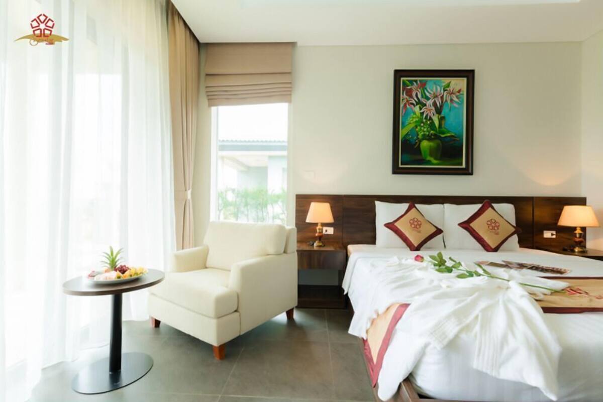 A hotel room with a bed, a chair, a small table with fruit, a painting on the wall, and a window with curtains.