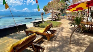 A beachside area with wooden lounge chairs and umbrellas, a flagpole with multiple flags, and a view of the sea and mountains in the background.