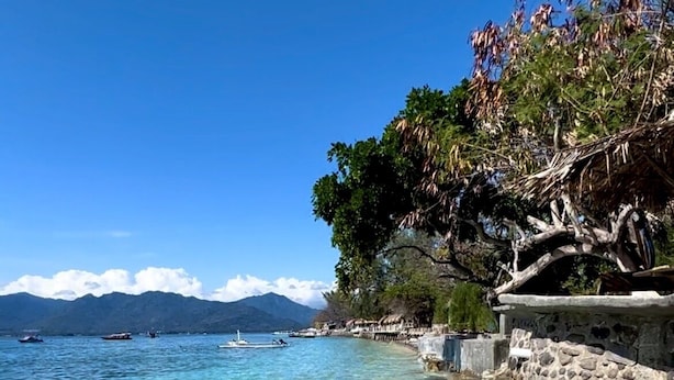 A clear coastal beach with calm waters, a stone wall, and a mountain range in the background.