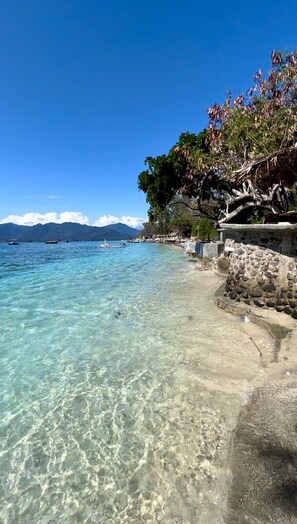 A clear coastal beach with calm waters, a stone wall, and a mountain range in the background. - Villa Bulan Madu Gili Air (Gili Air)