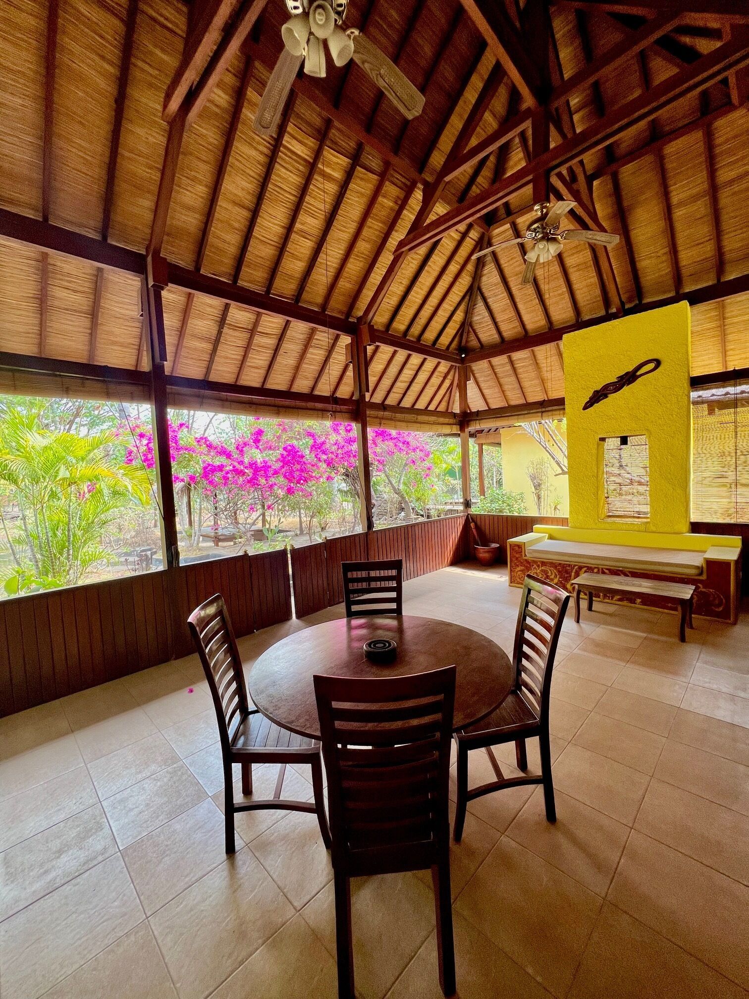 A covered outdoor dining area with a round table and chairs, a yellow wall with a window, and a ceiling fan.