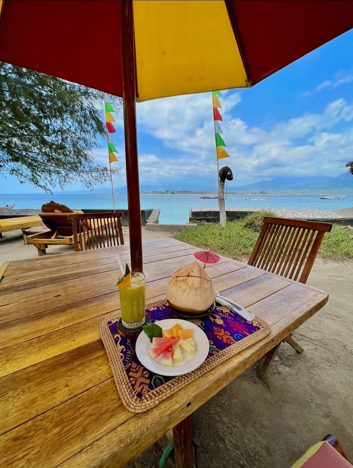Une table en bois surmontée d’un set de table en fibres tressées, une assiette de fruits, une noix de coco et une boisson, le tout placé sous un parasol rouge et jaune, sur une plage d’où l’on peut admirer la mer et les bateaux.