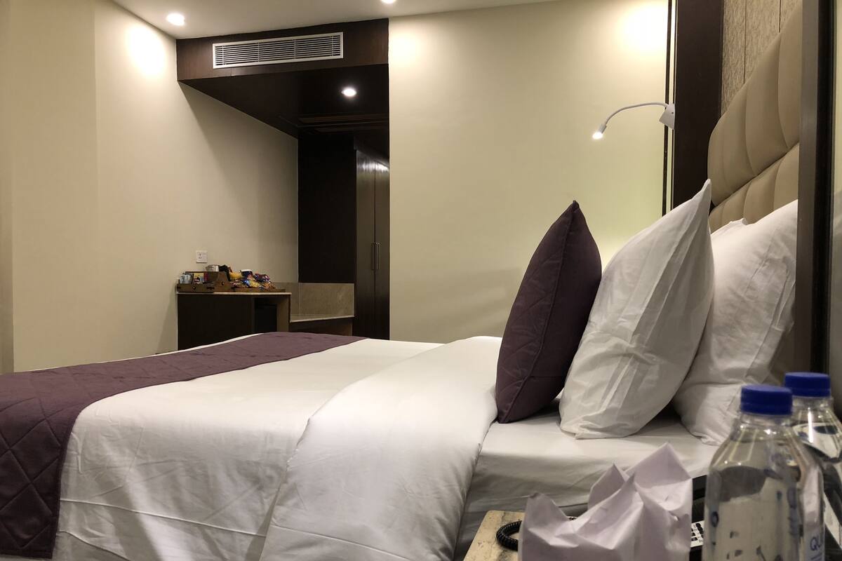 A hotel room with a bed, pillows, a nightstand, a lamp, and a water bottle.