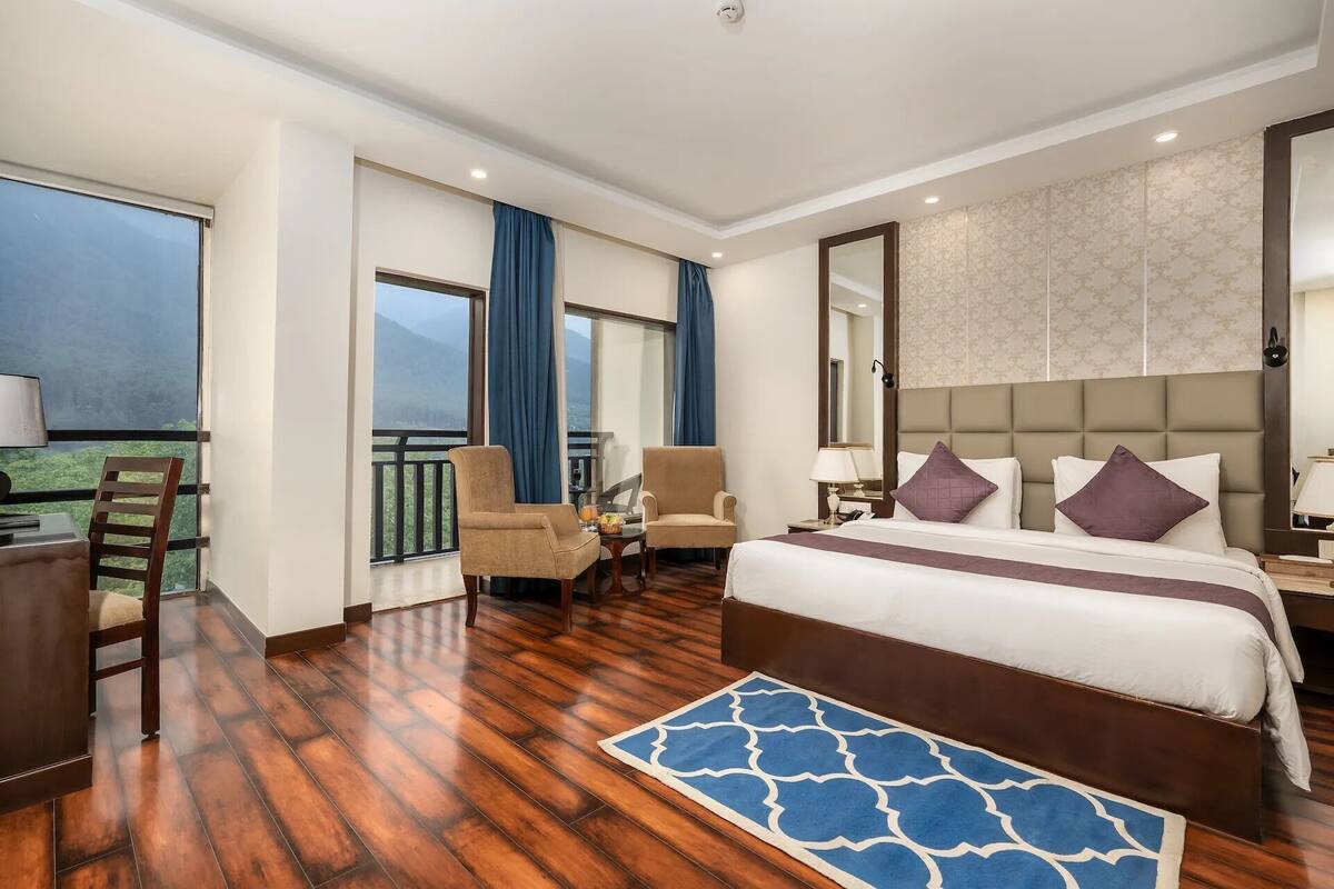 A hotel room with a large bed, two armchairs, a desk, a balcony with a view, and a patterned wall.