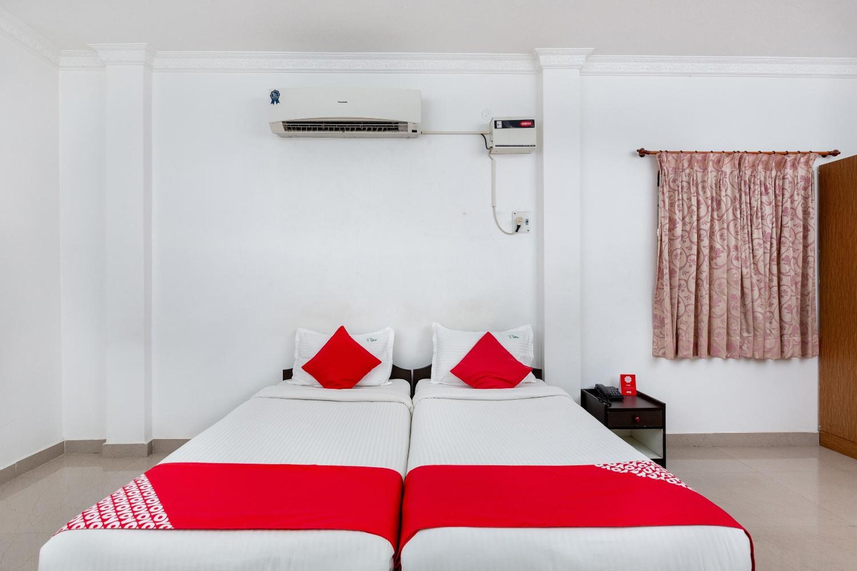 A hotel room with two single beds, a bedside table with a phone, a red and white bedspread, a wall-mounted air conditioner, and a patterned curtain.