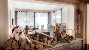 A cozy room with a wooden ceiling, a large window, a dining table with chairs, and a fireplace with logs and a teddy bear. - Logis Hôtel le Littoral (Evian-les-Bains)