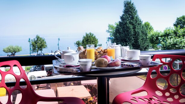 A balcony table with breakfast items, a view of the sea, and red chairs. - Logis Hôtel le Littoral (Evian-les-Bains)