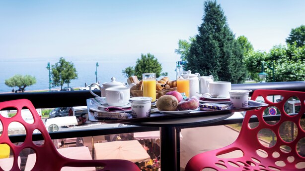 A balcony table with breakfast items, a view of the sea, and red chairs. - Logis Hôtel le Littoral (Evian-les-Bains)