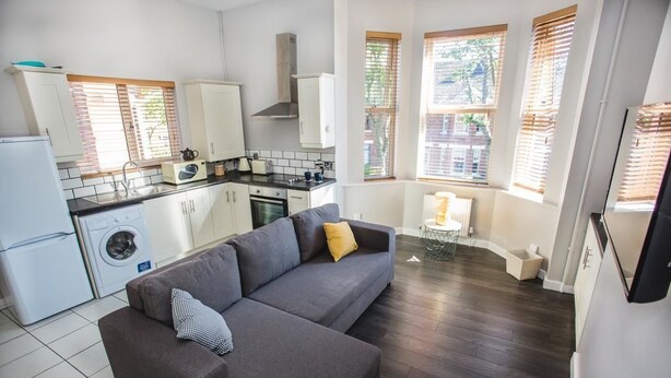 A compact kitchen with a built-in oven, a washing machine, and a dining area with a grey sofa and a small table. - Glandore Lodge (Belfast)