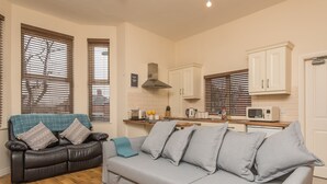A living room with a grey sofa and a black leather sofa, a kitchen area with white cabinets and a microwave, and large windows with blinds. - Glandore Lodge (Belfast)