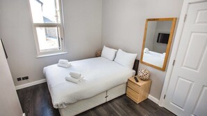 A neatly made bed with white linens and a set of towels, a bedside table with a remote control and decorative item, a large mirror, and a window with a view of the outside. - Glandore Lodge (Belfast)