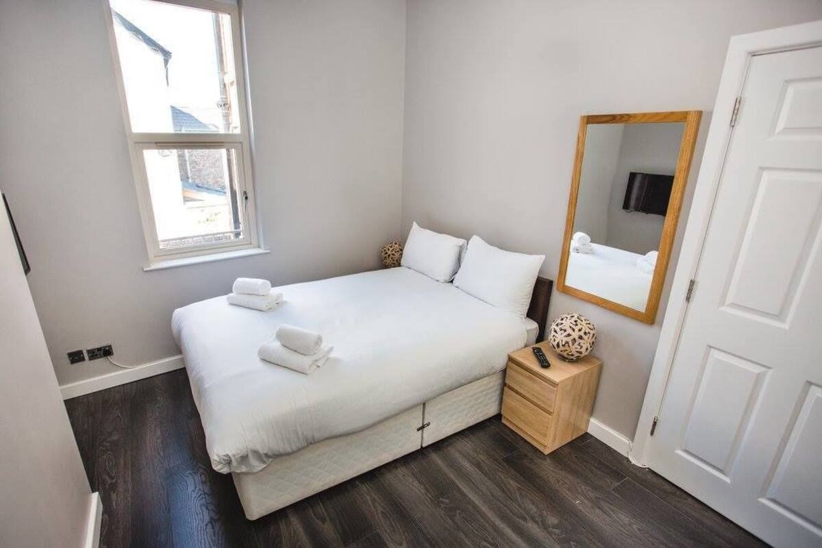 Un lit bien fait, avec du linge de lit blanc et un ensemble de serviettes, une table de chevet avec une télécommande et un objet décoratif, un grand miroir et une fenêtre donnant sur l’extérieur.