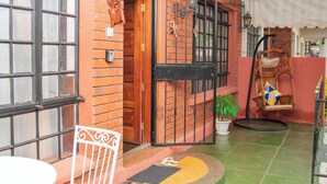 Un patio con pared de ladrillo, puerta de madera, una silla blanca, planta en maceta y una silla colgante.