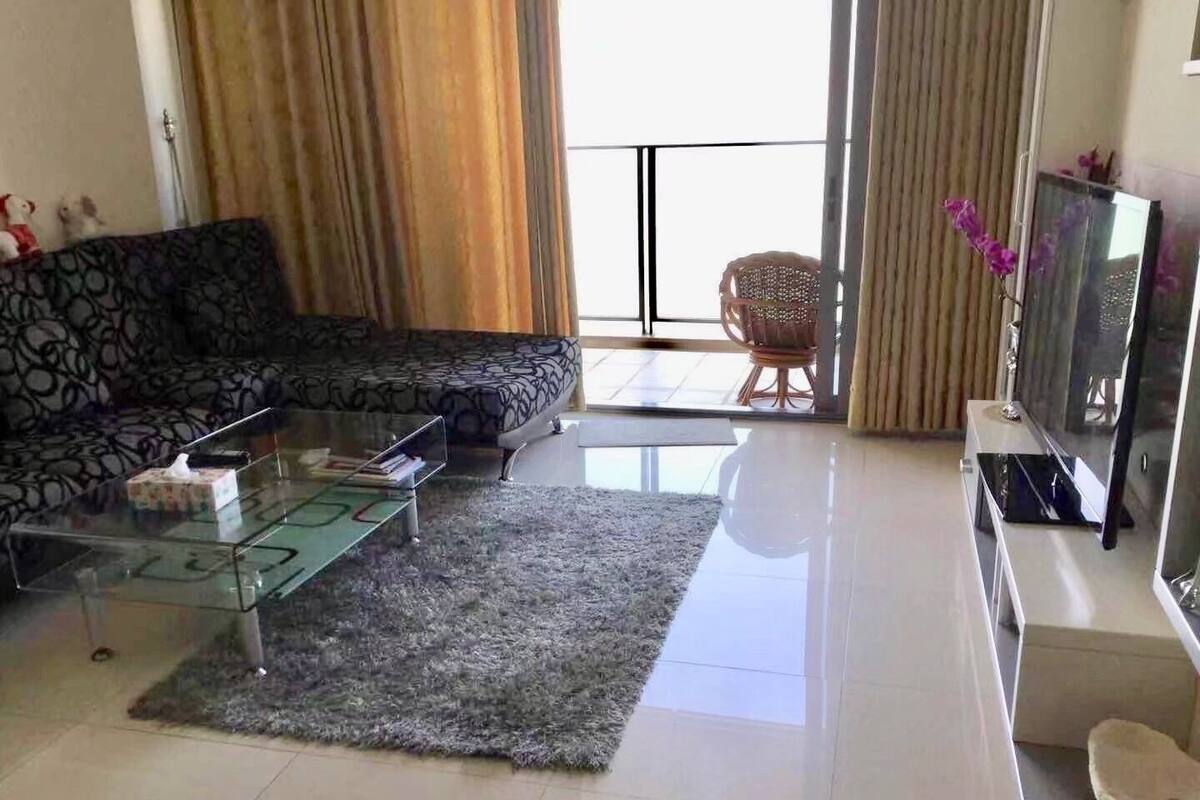 A modern living room with a glass coffee table, a patterned sofa, a television, and a wicker chair.