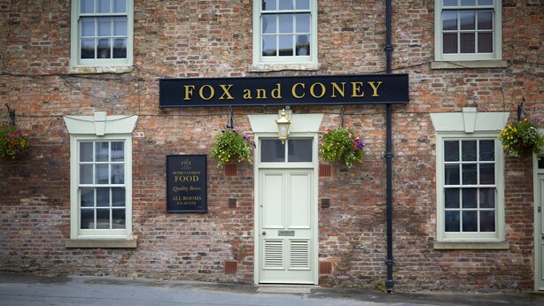Un bâtiment en briques sur lequel on peut lire l’inscription « FOX and CONEY », et en dessous, une plus petite pancarte indiquant : « NOURRITURE FAITE MAISON, bières de qualité, toutes les chambres sont équipées d’une salle de bain privée. »
