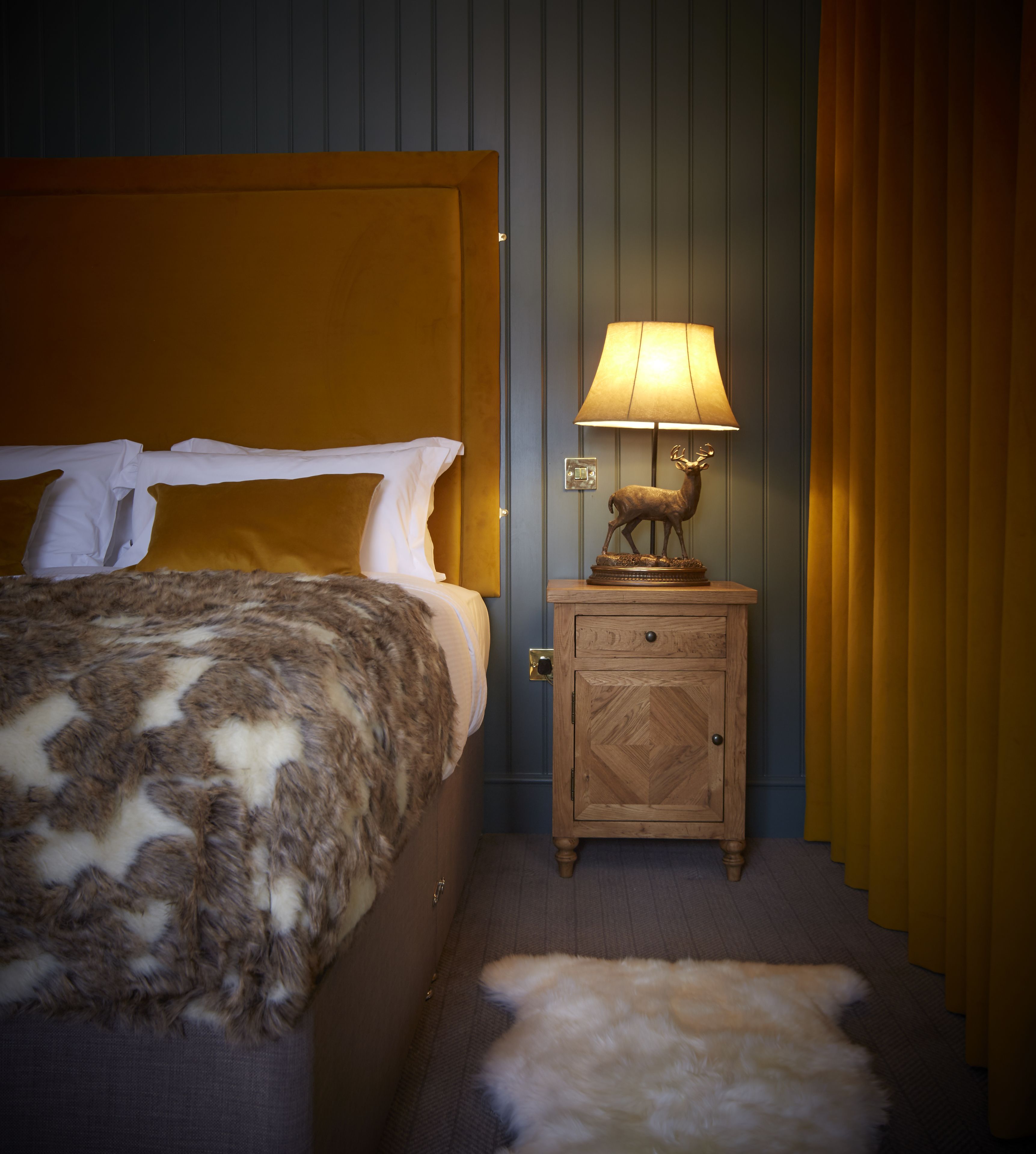 A bed with a patterned blanket, a nightstand with a lamp and a deer statue, and a fur rug on the floor.